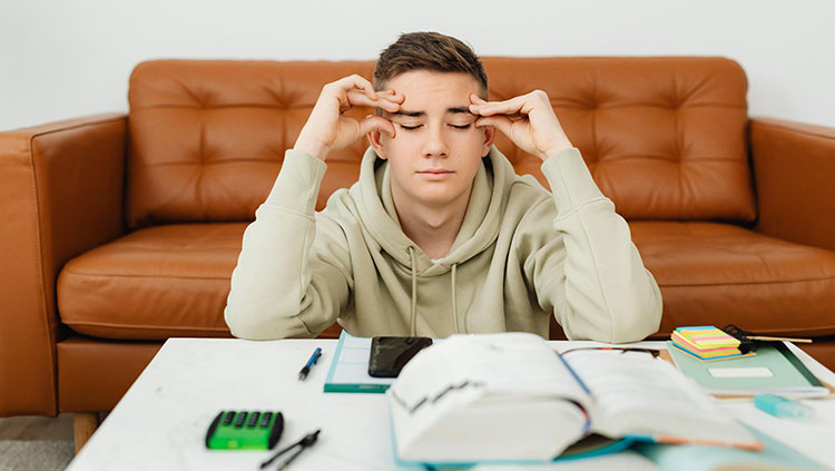 Teenager sitting on the floor in front of a couch, looking tired with eyes closed and fingers on temples, surrounded by open book, phone, and study materials.
